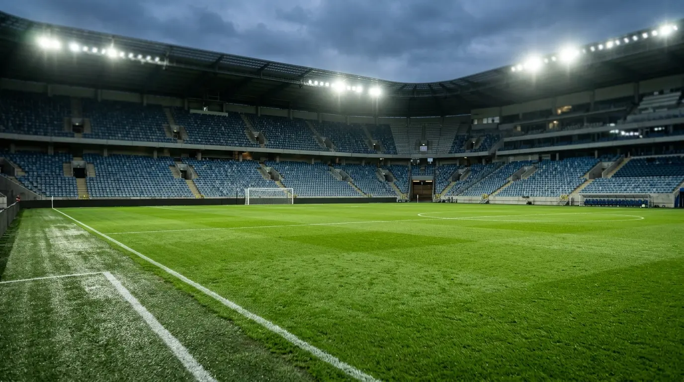 Leeres Fußballstadion am Abend mit beleuchtetem Rasen und eingeschaltetem Flutlicht vor unbesetzten Rängen.