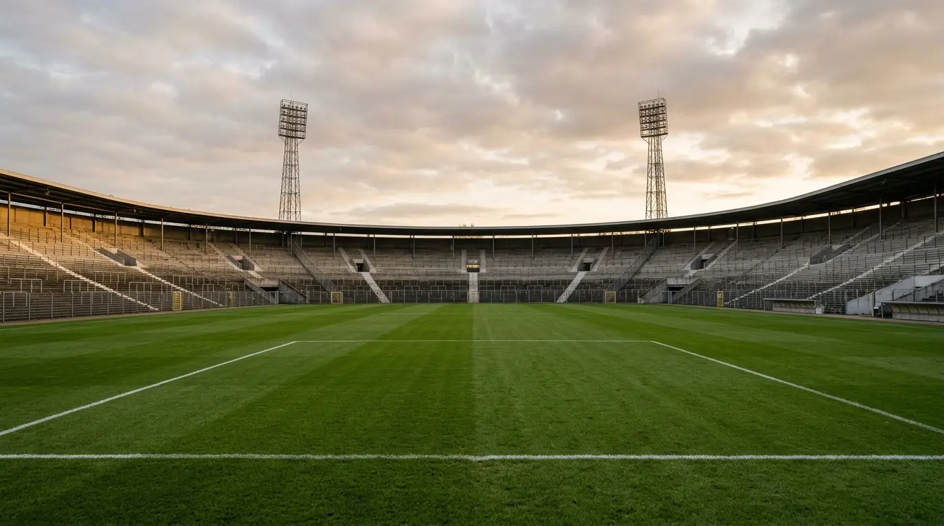 Leeres Fußballstadion mit gepflegtem Rasen im Vordergrund, Stehrängen und hohen Flutlichtmasten im Abendlicht.