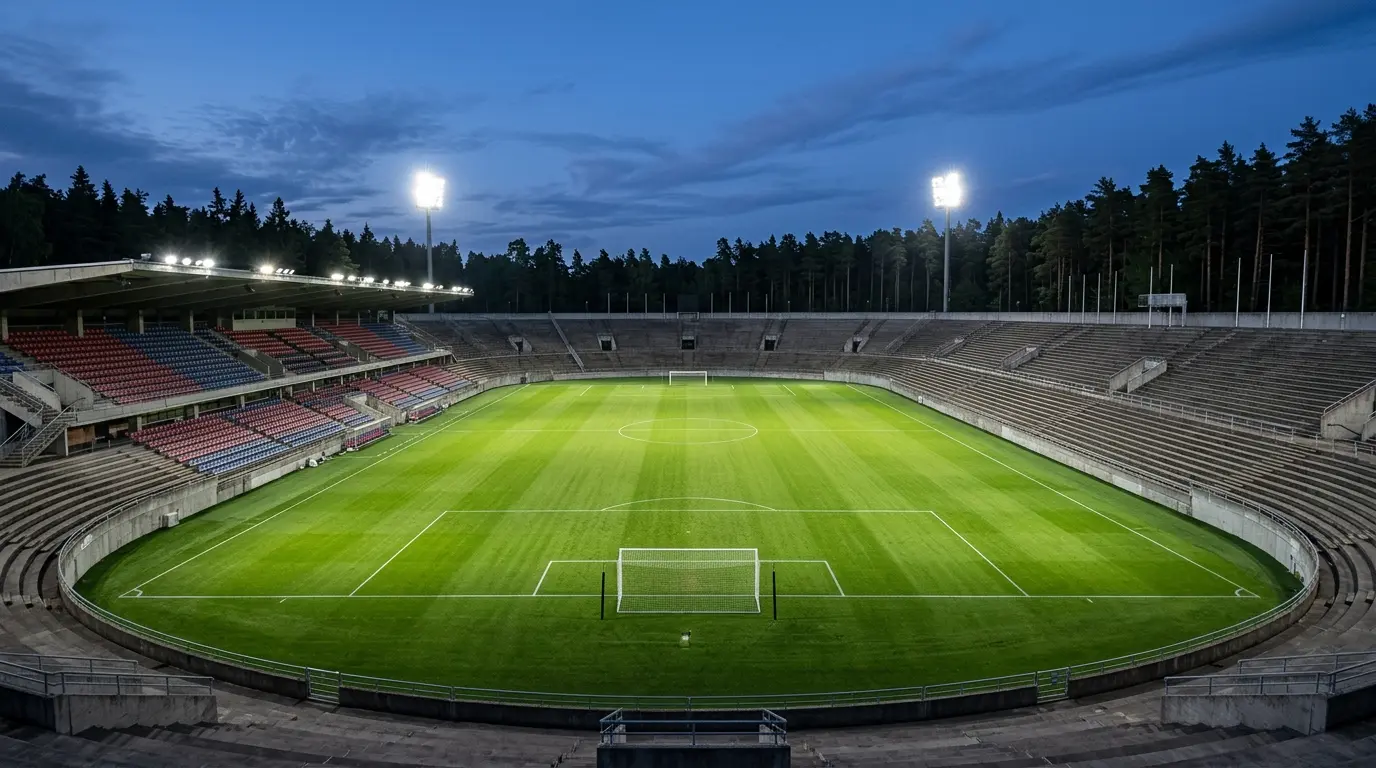 Ein leeres Fußballstadion bei Dämmerung mit erleuchtetem grünem Rasen und unbesetzten Stehplatztribünen.
