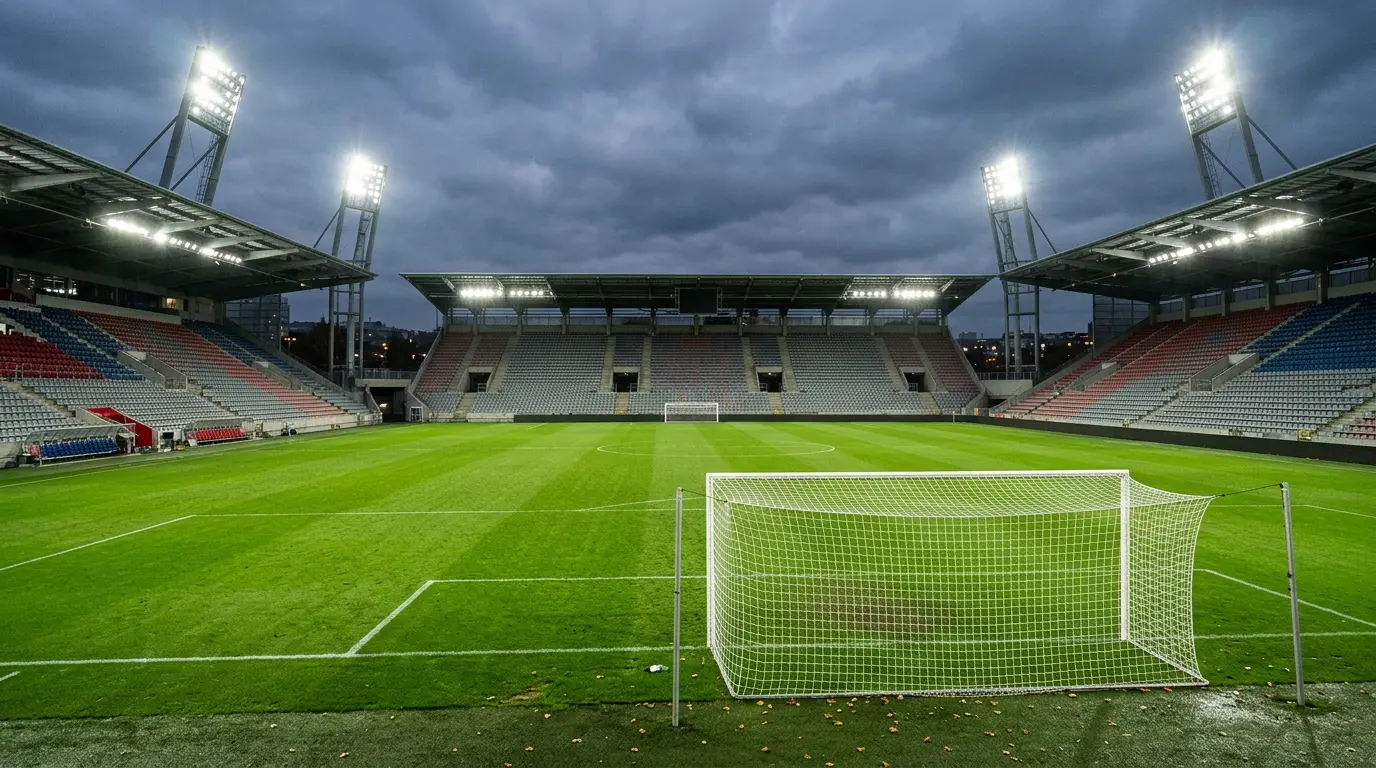 Leeres Fußballstadion am Abend mit grünem Rasen, weißem Tor und leuchtendem Flutlicht vor unbesetzten Rängen.