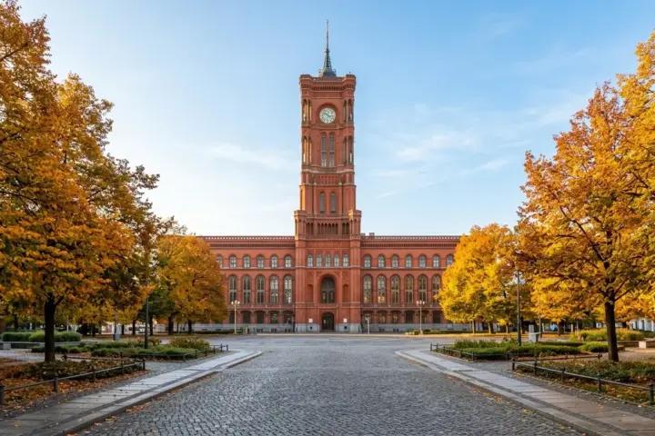 Die rote Klinkerfassade des Berliner Rathauses im warmen Herbstlicht hinter Bäumen mit gelben Blättern ohne Passanten.